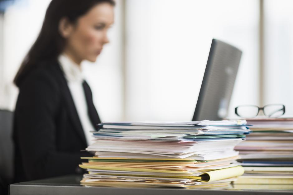 Ženska sedi za računalnikom ob kupu papirjev | Avtor: Profimedia