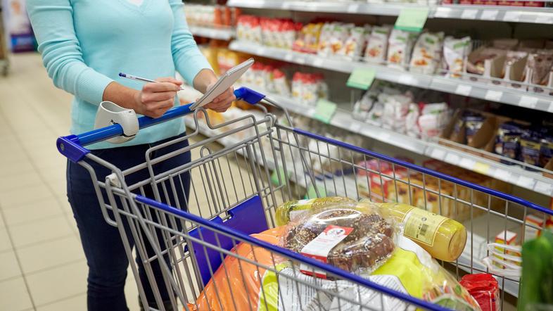 ženska v trgovini v roki drži nakupovalni seznam in ima pred seboj nakupovalni voziček